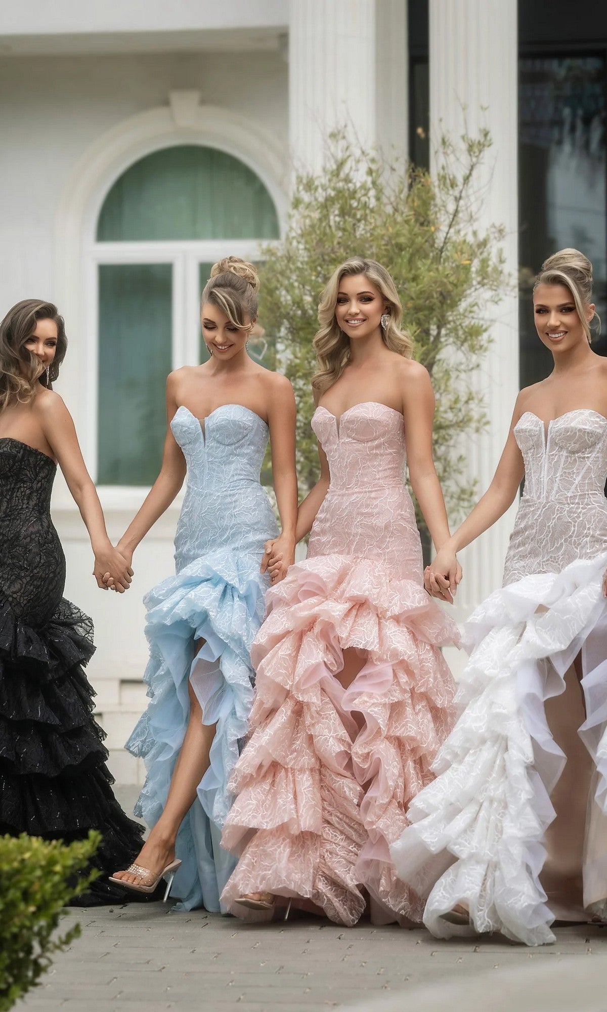Four women stand outdoors holding hands, each wearing the Long Formal Dress: Portia and Scarlett PS25250 in strapless styles—some with ruffled tiered skirts or boned corset bodices—in black, light blue, blush pink, and white. A columned white building is behind them.